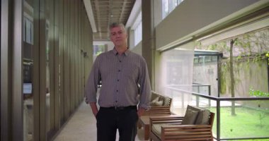 Orta yaşlı bir adam ofis binasının koridorunda güvenle duruyor hafif bir gülümsemeyle sakin ve profesyonel çevrede ulaşılabilir bir ifadeyle.