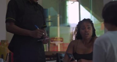 Afrika kökenli Latin garson masanın yanında oturuyor. Oturan kadınlardan sipariş alıyor. Bir misafirin eline telefon alıp dikkatle bakıyor.