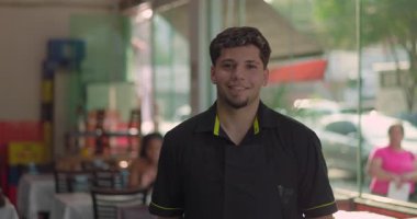 Gülümseyen genç garson, park halindeki arabalar ve ağaçlarla birlikte restoran penceresinin yanında poz veriyor. Gün ışığında neşeli ve rahat bir ifade takınıyor.