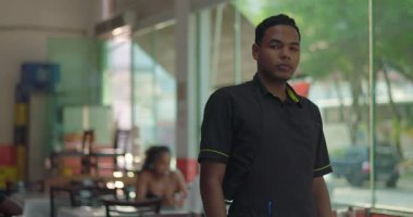 Genç erkek garson güvenle restoran penceresinin yanında duruyor, kollarını kavuşturmuş, müşterilerle ciddi bir yüz ifadesi ve camdan görülebilen bir sokak.