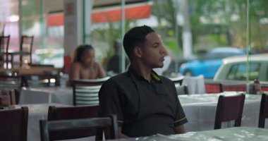 Yemek odasındaki pencerenin yanında oturan düşünceli genç garsonun, işteki sakin bir anında içsel bir ifadeyle dışarı bakışı.