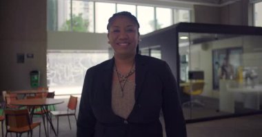 Kolları çapraz duran Afrikalı Amerikalı bir kadın modern ofis içinde güvenle gülümsüyor. Liderlik, profesyonellik ve yakınlık.