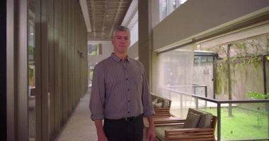 Orta yaşlı bir adam modern ofis alanının koridorunda duruyor. Kameraya tarafsız bir ifadeyle bakıyor. İş dünyası.