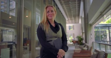 Koridorda duran bir kadın, göğsünü ve tabletini sıkıca tutuyor, iş ortamındaki gergin anlarda rahatsızlık veya duygusal hassasiyet gösteriyor.