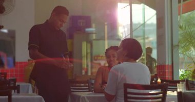 Restoran masasında oturan iki kadın akıllı telefondaki bir şeye tepki veriyor, gülümsüyor ve kapalı alanda camdan görülebilen tropikal bitkilerle el kol hareketi yapıyor.