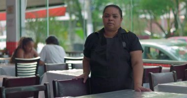 Tarafsız bir ifadeyle yemek alanında duran kadın restoran çalışanı, arka planda oturan ve penceresi olan sıradan bir oturma düzeninde sandalyeye uzanmış.