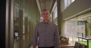 Orta yaşlı bir adam koridorda duruyor. Yumuşak bir gülümsemeyle ofisin iş yeri koridorunda geniş pencereli rahatlamış profesyonel bir ifadeyle.