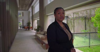 Afro-Amerikalı iş kadını kameraya bakıyor, sakin, modern bir koridorda duruyor bitkilerle ve camdan içeri doğal ışık salıyor.