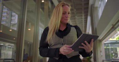 Modern koridorda nazik gülümsemeli tablet kullanan bir kadın, dijital ekranla etkileşimde bulunurken aynı zamanda tatmin olmuş ve işle ya da iletişimle meşgul görünüyor.