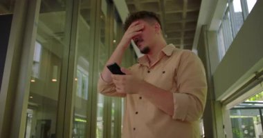Ofis koridorundaki genç adam akıllı telefona bakarken hayal kırıklığıyla tepki veriyor. Profesyonel ortamdaki hayal kırıklığına uğramış yüz ifadesi.