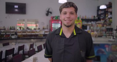 Gündelik restoranda kameraya bakan gülümseyen genç erkek garson, içecek dolapları ve açık büfe arka planda bulanık, dostane ve ulaşılabilir ifade