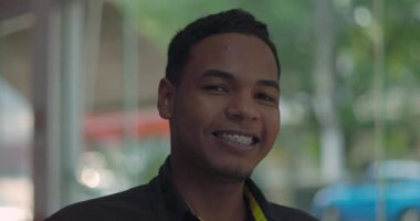 Gülümseyen genç garsona yakın çekim, kameraya bakarken neşeli yüz ifadesi, arka planda gün ışığı ve sokak ile restoranın içinde duran