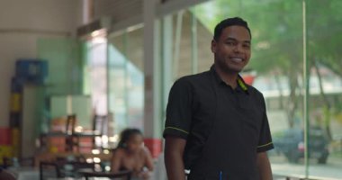 Gündelik yemek alanında pencerenin yanında duran gülümseyen genç erkek restoran çalışanı, kapalı oturaklı neşeli bir ifade ve arka planda görünen şehir sokağı