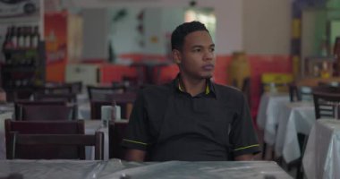 Genç restoran çalışanı boş yemek alanında oturuyor, pencerenin yanındaki sakin ve yansıtıcı duruşla mesafeye bakıyor.