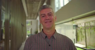 Orta yaşlı bir adam kameraya bakıyor. Profesyonel ofis koridorunu kapatıyor. Kendine güvenen ve sakin bir iş ifadesi sergiliyor.