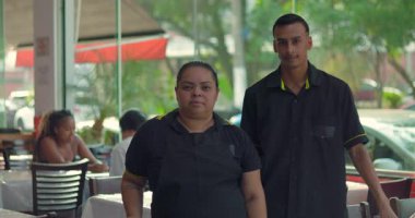 Lokantada genç erkek garsonun yanında duran Latin garson kolları yan yana rahatlamış, tarafsız ifadelerle doğrudan kameraya bakıyor.