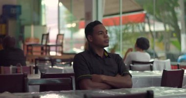 Genç bir restoran çalışanı kollarını kavuşturup yalnız başına oturuyor ve vardiyadan sonra neredeyse boş olan yemek odasında ciddi ve düşünceli bir tavırla ileri bakıyor.