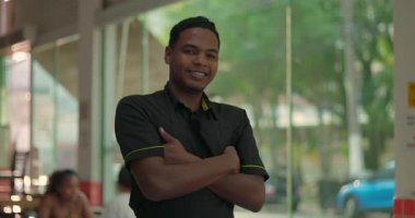 Pencere kenarında kollarını kavuşturarak poz veren gülümseyen genç bir restoran çalışanı, dışarıda sokak olan kapalı bir yemek salonunda neşeli bir ifade.