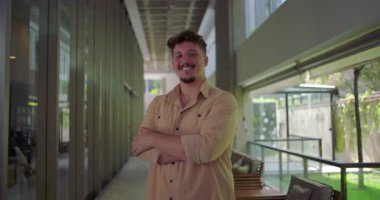 Ofis koridorunda kolları kavuşturulmuş, rahat ve olumlu bir şekilde gülen profesyonel iş ortamında rahat ve kendinden emin bir şekilde duran genç bir adamın vücut portresi.