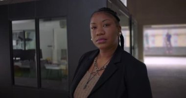 Cam ofis duvarının yanında duran Afro-Amerikalı kadın ciddi bir ifadeyle modern iş ortamında sakin ve kararlı görünüyor.