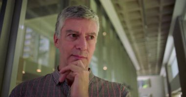 Orta yaşlı bir adam, modern ofis koridorunda kafa kafaya verip düşünceli bir poz veriyor. Karar verme ve derin düşüncelere dalmayı yansıtıyor.