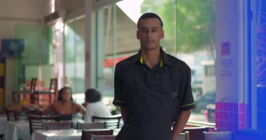 Camdan duvarın yanında duran genç bir restoran çalışanı. Elleri arkada. Arka planda gün ışığı ve sokak olan ciddi bir ifade.