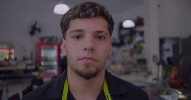 Ciddi genç bir restoran çalışanı kameraya tarafsız bir ifadeyle bakıyor. Arka planda görünen sıradan bir restoranın içi.