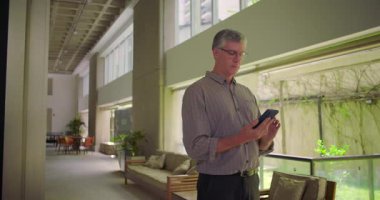 Orta yaşlı bir adam ofis koridorunda duruyor telefonu kulağına dayayıp modern kurumsal çevrede mesleki bir konuşma ya da iş görüşmesi yapıyor.