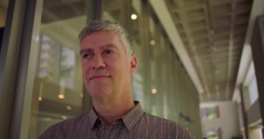 Orta yaşlı bir adam modern ofis koridorunda kurnaz bir gülümsemeyle profesyonel çevrede düşünceli, rahat ve iyimser bir ifadeyle yan yana bakıyor.