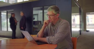 Orta yaşlı bir adam endişeli bir ifadeyle dizüstü bilgisayara bakıyor, ortak çalışma alanında masada oturuyor, elleri çenesinde, iş günü boyunca endişeli ve dalgın görünüyor.