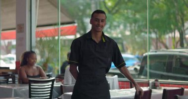 Gülümseyen genç bir restoran çalışanı gündelik yemek restoranının penceresinden sandalyeye yaslanıyor. Arka planda müşterileri olan neşeli bir ifade.