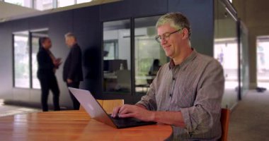 Orta yaşlı bir adam laptopta yumuşak bir gülümsemeyle çalışıyor, modern çalışma ortamında yuvarlak masada oturuyor, arka planda iş arkadaşlarının konuştuğu bir göreve odaklanıyor