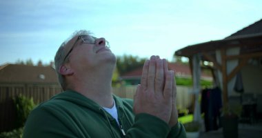 Elleri dua pozisyonunda, huzurlu ve içten bakan bir adam, arka planda bahçe ve yerleşim evleriyle donatılmış, sakin ve ruhsal bir ortam.