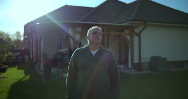 Dışarıdan bakan bir adam, arka planda bir ev, yeşil çimenler, huzurlu bir banliyö mahallesi, düşünceli bir ifade.