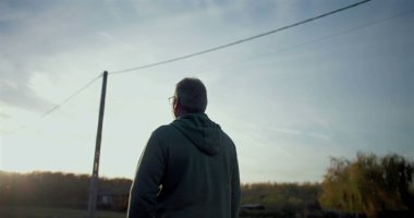 Kırsal bir yolda yürüyen bir adam, düşünceli bir şekilde ileriye bakıyor, sakin, açık bir çevre, yol boyunca sıralanmış elektrik direkleri ve ağaçlar, sakin kırsal atmosfer.