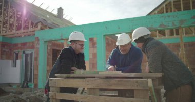 Planlar etrafında tartışan inşaat işçileri, sert şapkalar takanlar, konut inşaatının planlarını gözden geçirenler, arka planda görünen çerçeve ve duvarcılık