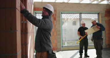 Tuğla duvarı inceleyen inşaat işçisi, arka planda iki işçi daha büyük pencereleri ve tamamlanmamış duvarları olan inşaat alanının planlarını inceliyor.