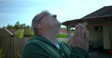 Elleri dua pozisyonunda, huzurlu ve içten bakan bir adam, arka planda bahçe ve yerleşim evleriyle donatılmış, sakin ve ruhsal bir ortam.