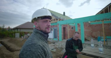 Ön planda güvenlik kaskı olan inşaat işçisi, gözlem alanı gelişimi, arka planda çalışan meslektaşı, kısmen inşa edilmiş ev yapısı görünür.