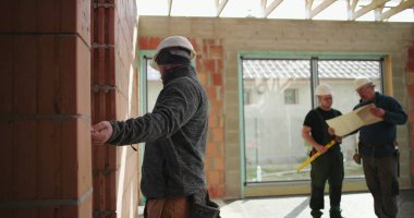 Tuğla duvarı inceleyen inşaat işçisi, arka planda iki işçi daha büyük pencereleri ve tamamlanmamış duvarları olan inşaat alanının planlarını inceliyor.