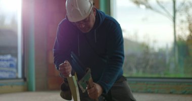 İnşaat işçisi odun parçalarını odaklama ve kuvvetle çekiçliyor, iç inşaat sahasında diz çöküyor, malzemeleri güvence altına alıyor, inşaat projesi devam ediyor.