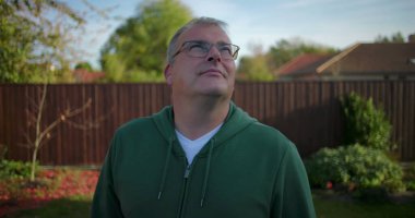 Arka bahçeye bakan bir adam, ahşap çitli ve yeşillikli bir banliyö ortamı, düşünceli bir ifade, sakin bir açık hava anı, yerleşim bölgesi.