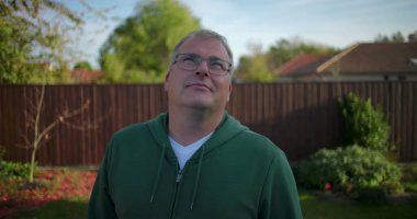 Arka bahçeye bakan bir adam, ahşap çitli ve yeşillikli bir banliyö ortamı, düşünceli bir ifade, sakin bir açık hava anı, yerleşim bölgesi.