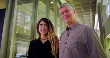 Erkek ve kadın ofis koridorundaki kameraya gülümsüyor, profesyonel çalışma alanındaki iş arkadaşları gibi yakın duruyorlar, rahat ve kendinden emin ifadeler.