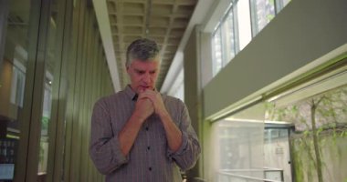 Orta yaşlı bir adam ofis koridorunda iki elini de yan yana tutarak aşağıya bakıyor. Kurumsal çevrede duygusal endişe ve endişe gösteriyor.