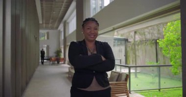 Ofis koridorunda kollarını kavuşturarak gülümseyen Afro-Amerikalı kadın cam pencereler ve koridorda yürüyen uzak figürler sergiliyor.