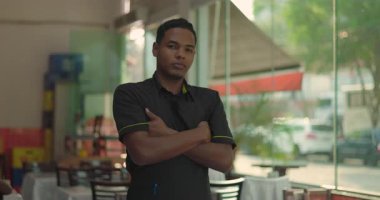 Lokanta çalışanı, kollarını pencereye yakın bir yerde, açık havada yemek yiyor, kendinden emin ve sakin ifade ediyor, arka planda doğal gün ışığı var.