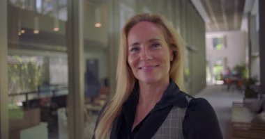 Koridorda dururken doğal bir şekilde gülümseyen kadın, kameraya yaklaşılabilirlik ve modern kapalı mekan ortamında profesyonel varlıkla bakıyor.