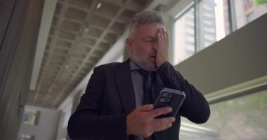 Orta yaşlı iş adamı gözlerini kapatır ve elleriyle yüzüne dokunur. Modern ofis koridorunda doğal ışık alan akıllı telefona bakarken şaşkına döner.