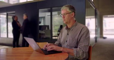 Orta yaşlı bir adamın açık ofis ortamında dizüstü bilgisayarda yazması, ciddi ve odaklanmış bir ifade, meslektaşları arka plandaki sohbete katılırken işine devam etmesi.
